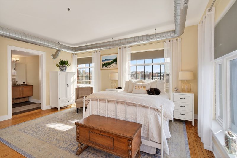 Bright primary bedroom with windows on two walls, exposed ductwork, hardwood floors and an en-suite bath through the door