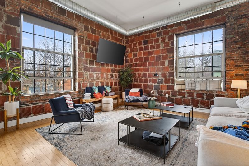Corner living room with original red brick walls, two tall factory windows, exposed ductwork and hardwood floors
