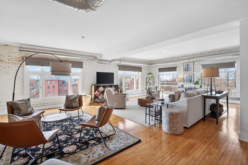 Sunlit corner living room with white-painted brick, wide factory windows looking over Allentown rooftops, and hardwood floors