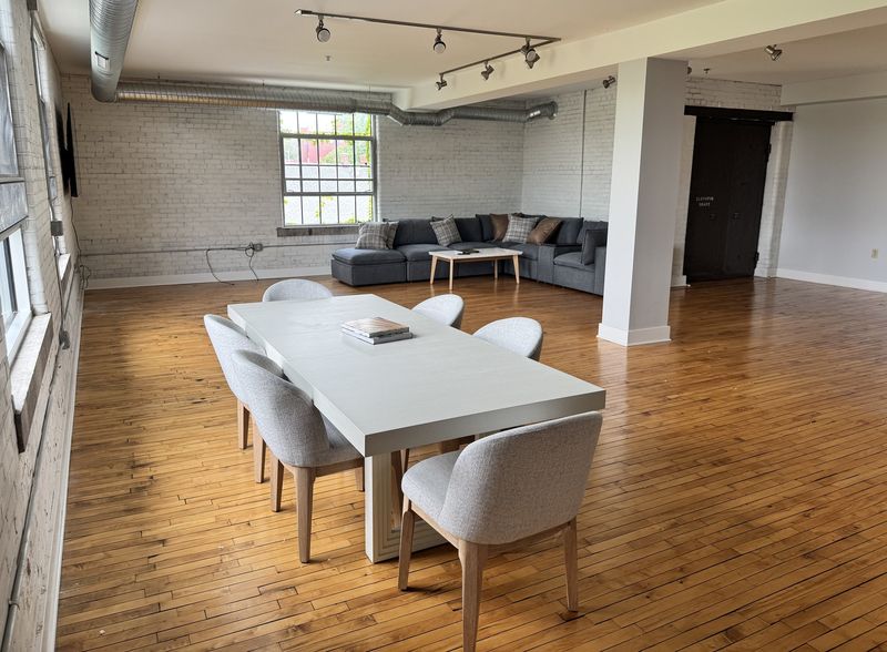 Wide open loft with original strip hardwood flooring, white-painted brick, exposed ductwork and track lighting, with a dining table and sectional