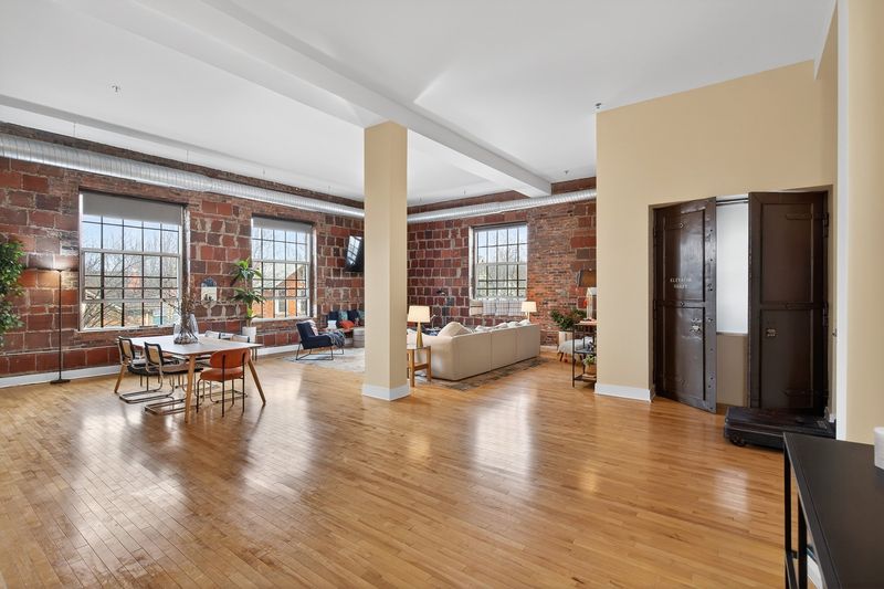 An open loft seen end to end: hardwood floors, a structural column, red brick walls and a run of factory windows